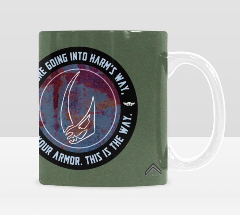 Pode incluir: Uma caneca de cer&acirc;mica verde com uma al&ccedil;a branca. A caneca apresenta um design circular com uma caveira com chifres branca e o texto "WE'RE GOING INTO HARM'S WAY. PUT ON YOUR ARMOR. THIS IS THE WAY." O design tem um aspeto envelhecido.