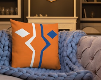 May include: Orange cushion with a blue and white geometric design. The cushion is on a blue knitted throw blanket.