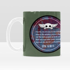 Pode incluir: Caneca de cer&acirc;mica verde com uma al&ccedil;a branca. A caneca apresenta um design circular com um personagem de desenho animado e texto de Ef&eacute;sios 6:10-11. O design tem um aspecto enferrujado e desgastado.