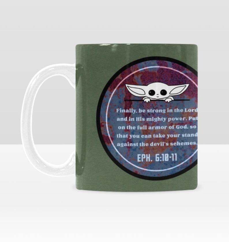 Pode incluir: Caneca de cer&acirc;mica verde com uma al&ccedil;a branca. A caneca apresenta um design circular com um personagem de desenho animado e texto de Ef&eacute;sios 6:10-11. O design tem um aspecto enferrujado e desgastado.
