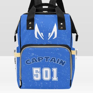 Könnte beinhalten: Blauer Rucksack mit weißen Akzenten und dem Text "CAPTAIN 501". Der Rucksack hat schwarze Träger und ein weißes Emblem auf der Oberseite. Die Tasche hat ein strukturiertes Muster und goldene Reißverschlüsse.