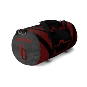 Könnte beinhalten: Rote und schwarze Reisetasche mit einem stilisierten "D" an der Seite. Die Tasche hat einen schwarzen Riemen und einen Reißverschluss. Der Text "YOUR NAME HERE" ist auf der Tasche aufgedruckt.