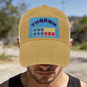 Könnte beinhalten: Senfgelbe Trucker-Cap mit einem blau-weißen Grafikdesign mit der Aufschrift "THRAWN". Die Grafik zeigt auch eine Reihe von blauen, gelben und roten Rechtecken. Die Kappe hat einen Mesh-Rücken und einen gebogenen Schirm.