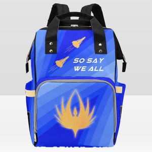 May include: Blue backpack with a front pocket featuring a gold emblem and the text "SO SAY WE ALL." The backpack has black straps and handles, with gold rocket ship graphics. The design is a mix of blue shades.
