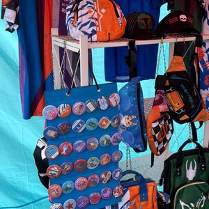 May include: A white metal rack displaying a variety of items for sale, including a blue and white backpack with the text "I am no Jedi", a green backpack with the text "SPECTRE", a blue and white bag with a chequered pattern, a blue bag with a moon and stars pattern, a blue bag with a variety of colourful buttons, and two blue and white shirts with the text "REX" and "ONCE A REBEL ALWAYS A REBEL".