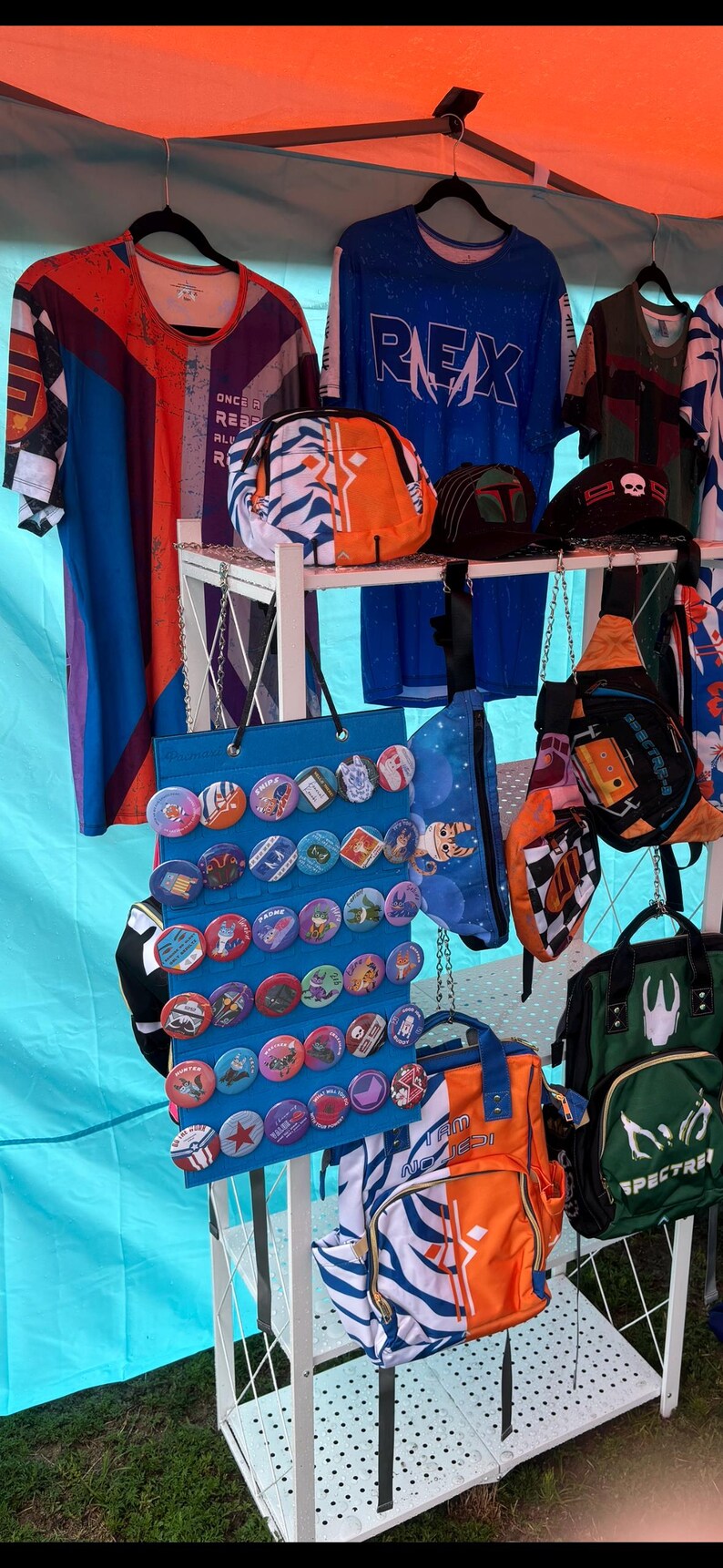 May include: A white metal rack displaying a variety of items for sale, including a blue and white backpack with the text "I am no Jedi", a green backpack with the text "SPECTRE", a blue and white bag with a chequered pattern, a blue bag with a moon and stars pattern, a blue bag with a variety of colourful buttons, and two blue and white shirts with the text "REX" and "ONCE A REBEL ALWAYS A REBEL".