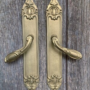 May include: Two gold-colored door handles with ornate, vintage-style detailing. Each handle has a long, rectangular backplate with decorative flourishes at the top and bottom, and a curved handle. The handles are set against a wooden background.