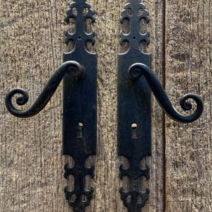 May include: A pair of black, decorative door handles with a gothic design. Each handle features a scrolled lever and an ornate backplate with cut-out patterns. The handles are mounted on a weathered wooden surface.