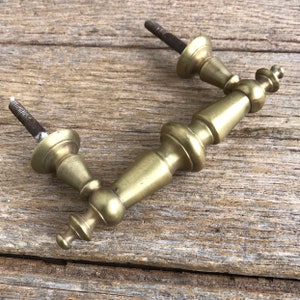 Puede incluir: Un tirador de cajón de latón vintage con un diseño curvo. El tirador presenta una forma ornamentada con dos postes roscados para el montaje. El latón tiene una pátina ligeramente envejecida, lo que sugiere su origen vintage. Herrajes para la restauración de muebles.