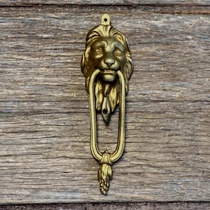 May include: A brass door knocker featuring a sculpted lion head with a flowing mane and a long, oval-shaped ring. The lion head has detailed facial features, and the bottom of the ring terminates in a decorative, textured leaf-like finial.