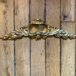 Può includere: Un frontone decorativo da parete color oro caratterizzato da un design ornamentale e simmetrico con motivi a foglia intagliati, volute e un elemento centrale a forma di conchiglia. L'oggetto presenta una finitura metallica scura.