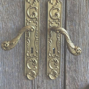 May include: A pair of antique-style brass door handles with ornate, swirling scrollwork patterns on the rectangular backplates. Each handle features a curved lever design and a central keyhole cutout, finished in a matte, muted gold tone.