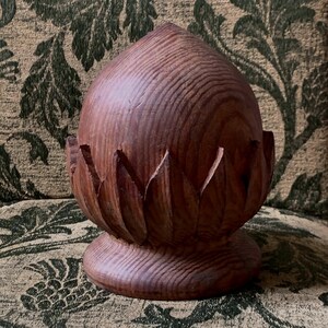 May include: A carved wooden finial featuring a rounded, pointed top section with a visible wood grain pattern. The base is circular and supports a ring of carved, petal-like segments that surround the lower portion of the central dome structure.