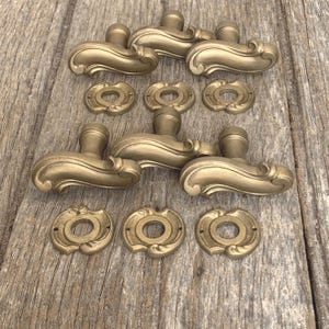 May include: A collection of antique brass door handles and matching backplates. The handles have a curved, ornate design, and the backplates are round with decorative edges. The hardware is arranged on a weathered wooden surface.