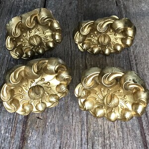 Könnte beinhalten: Vier goldfarbene Metall-Schubladengriffe mit floralem Design. Jeder Griff hat eine erhabene Mitte mit einem Wirbelmuster und eine kleine Perle in der Mitte.
