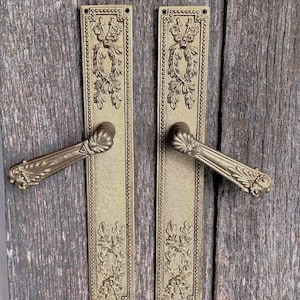 Puede incluir: Dos manijas de puerta de latón a juego con placas rectangulares. Cada placa presenta un borde de cuentas y motivos de guirnaldas florales en relieve en la parte superior e inferior. Las manijas tienen un diseño de concha en la base.