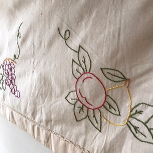 Könnte beinhalten: Cremefarbener bestickter Stoff mit einer Traube, einem Pfirsich und einer Zitrone. Die Stickerei verwendet grüne, violette, rote und gelbe Fäden. Der Stoff hat einen dekorativen, genähten Rand entlang der Unterkante.