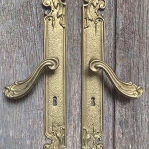 May include: A pair of gold-colored metal door handles with long, rectangular backplates. Each handle features an ornate, curved lever and intricate scrollwork detailing at the top and bottom ends. A small keyhole is centered on each backplate.