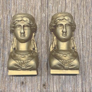 May include: A pair of identical gold-colored wall-mounted busts featuring a stylized human head with braided hair, a beaded necklace, and a draped garment. Each piece has a flat base and two small circular holes for mounting purposes.