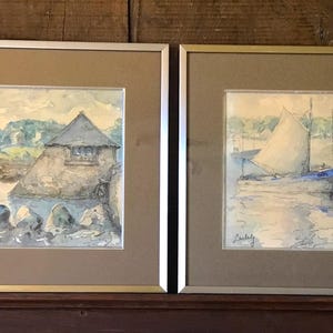 Könnte beinhalten: Zwei gerahmte Aquarelle. Das Bild links zeigt ein kleines Gebäude an einem felsigen Ufer mit Wasser im Vordergrund. Das Bild rechts zeigt zwei Segelboote in einem Hafen mit Wasser im Vordergrund.