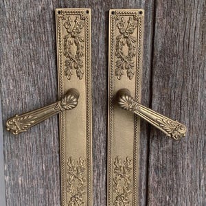 May include: Two antique brass door handles with ornate floral and leaf designs. The handles are rectangular with a long, narrow plate and a curved handle. The background is a weathered wooden surface.
