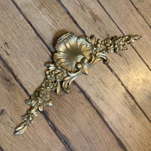 Könnte beinhalten: Eine goldfarbene dekorative Möbelapplikation mit einem zentralen Muschelmotiv, das von skulpturalen Blumen- und Blattdetails umgeben ist. Das Stück hat eine längliche, asymmetrische Form mit detaillierten Reliefschnitzereien auf der gesamten Oberfläche.