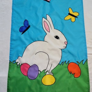 May include: A white bunny rabbit with pink ears sits on green grass with colorful Easter eggs. There are three butterflies in the sky, one blue, one pink, and one yellow.