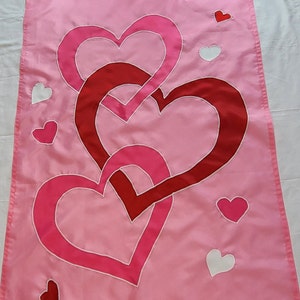 May include: Pink garden flag featuring a design of overlapping hearts in shades of pink and red. White hearts are scattered across the flag. The flag is made of a smooth, likely polyester material, and is designed for outdoor use.
