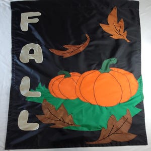May include: Black outdoor flag with the word "FALL" in cream-colored letters. The flag features two orange pumpkins, brown leaves, and green foliage. A festive autumn decoration.