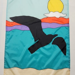 May include: A colorful flag with a black silhouette of a bird flying over a turquoise ocean with a yellow sun and white cloud in the sky. The ocean is layered with orange, purple, and blue stripes. The bottom of the flag is a tan beach.