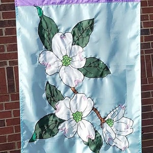 May include: A rectangular garden flag featuring a light blue background with a purple border at the top and bottom. The center displays a branch with three white dogwood flowers, each with a green center, surrounded by dark green leaves.
