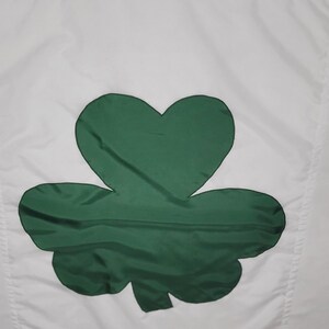 May include: White flag with a large green shamrock emblem. The shamrock is a three-leaf clover, a symbol of Ireland, and is centered on the white background. The flag has a simple, clean design.