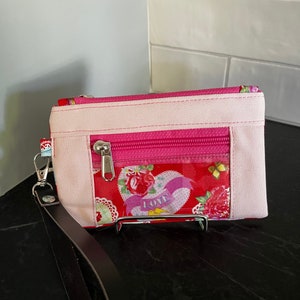 May include: A pink and red fabric zippered pouch with a clear plastic window featuring a heart with the word "LOVE" on it. The pouch has a black leather strap.
