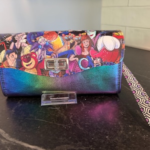 May include: A colorful wallet with a flap closure and wrist strap. The top of the wallet features a cartoon design with various characters. The bottom portion of the wallet has a gradient of blue, green, and purple. The wrist strap has a geometric pattern.