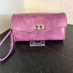 May include: A plum-colored clutch wallet with a textured finish and a gold-colored clasp. The wallet has a wrist strap and is displayed on a black surface. The design is simple and elegant.