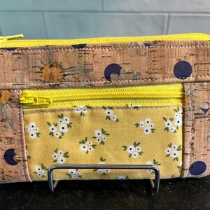 May include: A rectangular cork wallet with a yellow floral fabric panel and a yellow zipper. The cork has a natural wood grain pattern with dark blue polka dots. The fabric panel features small white flowers on a yellow background.
