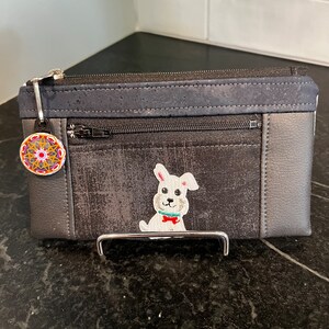 May include: A black and gray zippered pouch with a cork accent and a white embroidered dog with a red bow tie. The pouch has a small round charm with a colorful mandala design.