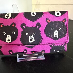 May include: A magenta wallet with a repeating pattern of black bear faces. The bears have white muzzles, black eyes, and red ears. The wallet is closed and has a snap closure.