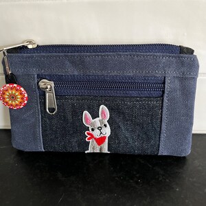 May include: A small blue zippered pouch with a white embroidered French bulldog wearing a red bandana. The pouch has a second zippered pocket on the front.