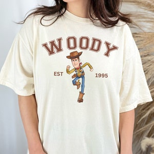 Puede incluir: Camiseta color crema con la palabra "WOODY" en letras marrones sobre una imagen de dibujos animados de Woody de Toy Story. El texto "EST 1995" est&aacute; debajo. La camiseta tiene un corte holgado y oversize.