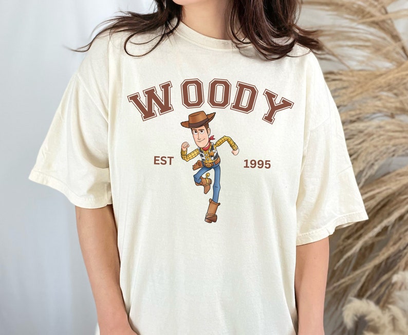 Puede incluir: Camiseta color crema con la palabra "WOODY" en letras marrones sobre una imagen de dibujos animados de Woody de Toy Story. El texto "EST 1995" est&aacute; debajo. La camiseta tiene un corte holgado y oversize.