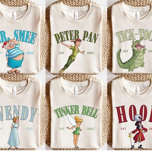 Op de afbeelding: Zes beige t-shirts met verschillende Peter Pan-karakters en tekst. De shirts tonen Mr. Smee, Peter Pan, Krokodil Tik-Tak, Wendy, Tinkerbell en Captain Hook. Elk shirt heeft de naam van het personage en "EST 1953" erop gedrukt.