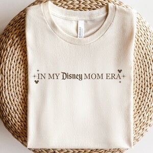 May include: A beige t-shirt with the text "IN MY DISNEY MOM ERA" in brown lettering with a Mickey Mouse silhouette and stars on either side.