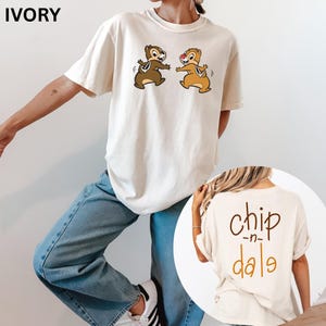 May include: Ivory-colored t-shirt featuring Chip and Dale cartoon characters. The front of the shirt displays the two chipmunks in a playful pose. The back of the shirt has the words "chip -n- dale" in a brown and orange color.