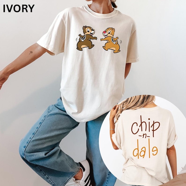 May include: Ivory-colored t-shirt featuring Chip and Dale cartoon characters. The front of the shirt displays the two chipmunks in a playful pose. The back of the shirt has the words "chip -n- dale" in a brown and orange color.