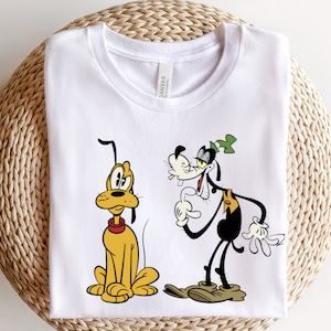 May include: White t-shirt with a cartoon image of Pluto and Goofy. Pluto is a yellow dog sitting down and Goofy is a black and white dog standing up.
