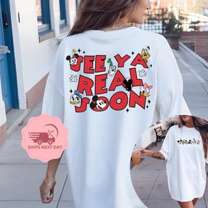 May include: White t-shirt with a red and black graphic print of Mickey Mouse and other Disney characters. The text on the shirt says "See Ya Real Soon".