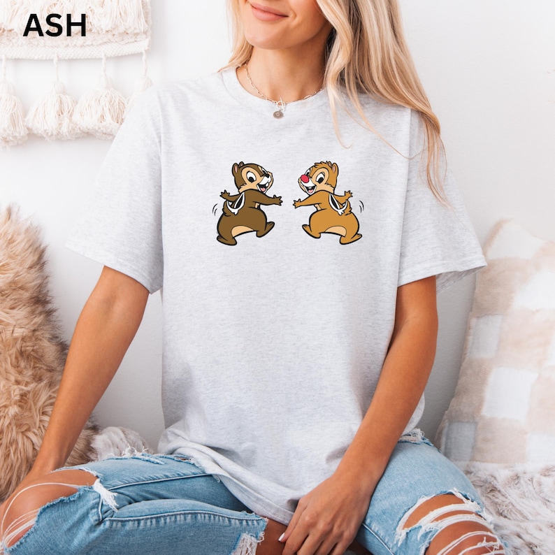 May include: Ash-colored t-shirt featuring a cartoon graphic of two chipmunks. The chipmunks are depicted in brown and tan with black outlines, arms outstretched, and smiling. The shirt is a light gray color.