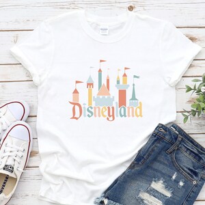 Op de afbeelding: Wit T-shirt met een Disneyland kasteelafbeelding in pastelkleuren en het woord "Disneyland". Het T-shirt wordt gecombineerd met een spijkerbroek en witte sneakers. De afbeelding is geplaatst tegen een houten achtergrond.