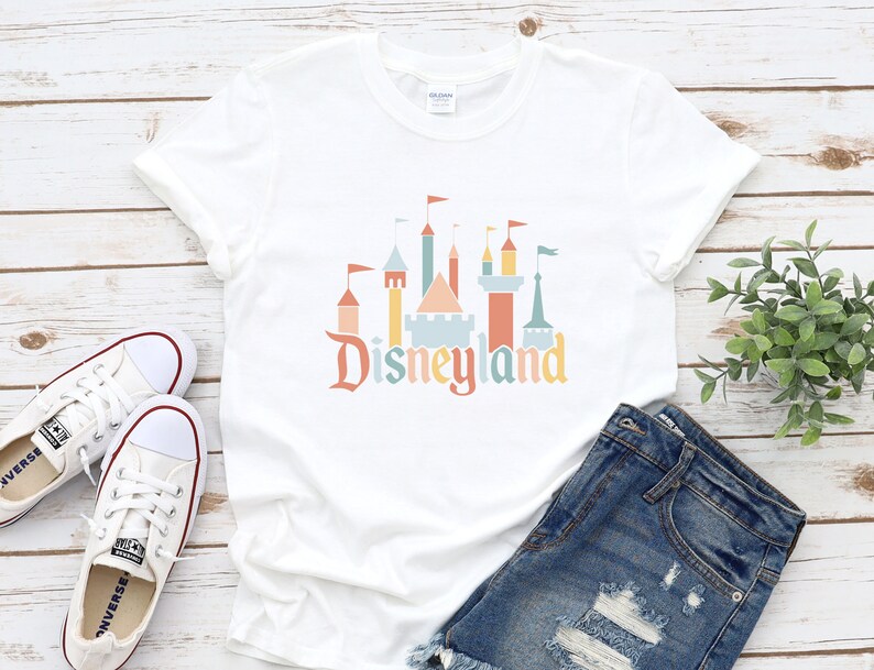 Op de afbeelding: Wit T-shirt met een Disneyland kasteelafbeelding in pastelkleuren en het woord "Disneyland". Het T-shirt wordt gecombineerd met een spijkerbroek en witte sneakers. De afbeelding is geplaatst tegen een houten achtergrond.